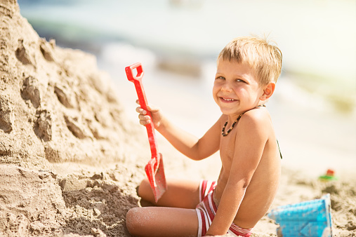 Little boy smiling into the camera, having fun building a sandcastle on the beach. He is holding a red sand shovel next to a big castle where he is sculpting windlows. The summer sun is flooding the beach with warm light.