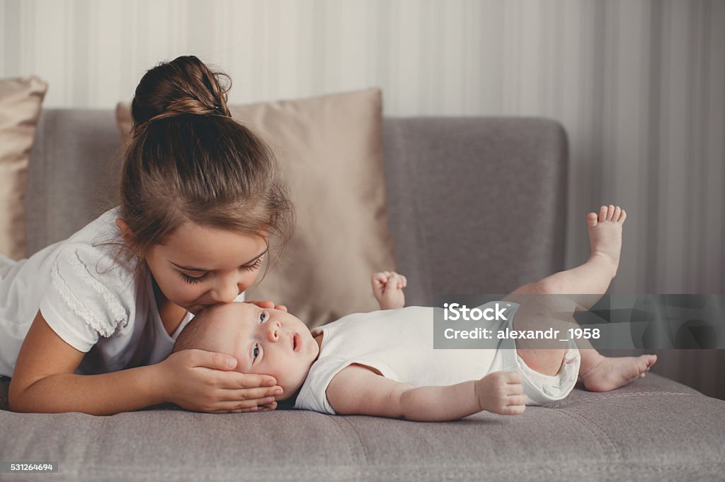 Little girl with a newborn baby brother A little girl five years old,a brunette,dressed in a white shirt and white pants,spends time together with her newborn brother,three-month-old boy,dressed in a white t-shirt and white panties,at home in the bedroom Sister Stock Photo