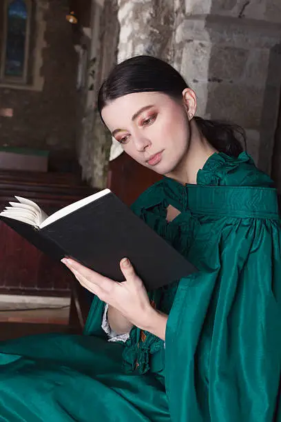 Dark-haired, attractive Victorian lady wearing a green dress and cloak, sitting in a church pew reading her prayerbook.