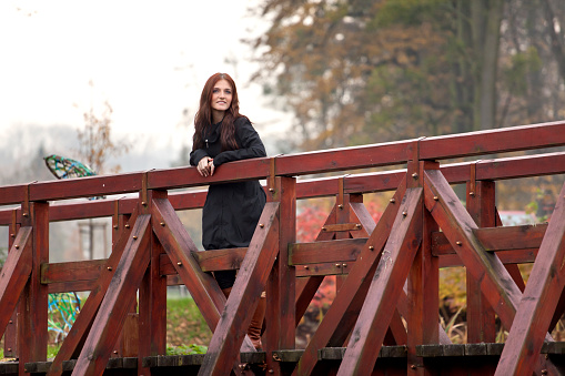 Attractive young brunette possing outdoors. She is standing on wood footbridge.in park is leaning on the railing, looking at the camera and laughing. Autumn atmosphere, selective focus.