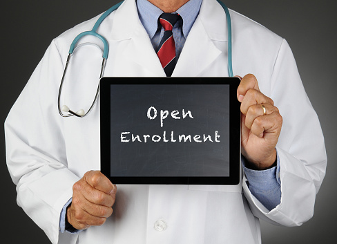 Closeup of a doctor holding a tablet computer with a chalkboard screen with the words Open Enrollment. Man is unrecognizable.