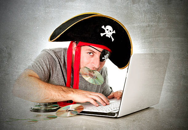 man in pirate hat downloading music and movies computer files man dressed as pirate with CD in his mouth at computer laptop downloading music files and movies in copyright violation and illegal internet piracy concept pirate dvds stock pictures, royalty-free photos & images