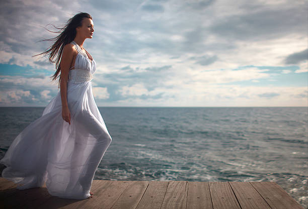 Gorgeous woman in white dress standing at pier close to cliffs. Ocean landscape view at windy day.