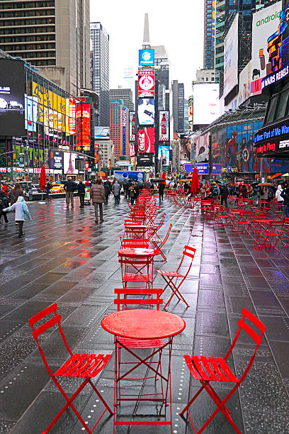 Unusual view of empty rain-soaked red tables and chairs on the paving at Times Square. Umbrellas and raincoats are essential as people are drawn to visit the landmark despite the bitter wintry weather.  Tables and chairs are main focus of image.  Lights reflect on the wet pavement.  Vertical.