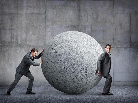 Two businessmen working against each other while trying to move a large sphere.