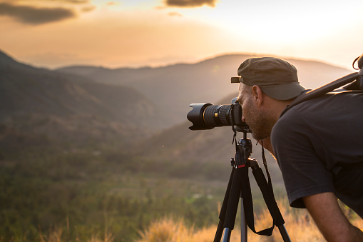 A matured man photographer is looking through the viewfinder and takes photographs of hills and mountains at sunset time, with a tele photo lens mounted on tripod.