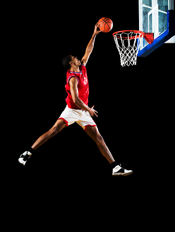 Young African American basketball player jumping high and slum dunking. Isolated on black.