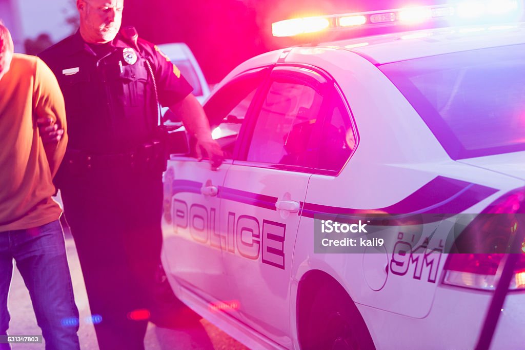 Police officer arresting a young man at night Police officer making an arrest, escorting a young man in handcuffs toward the back seat of his police car, at night. Arrest Stock Photo