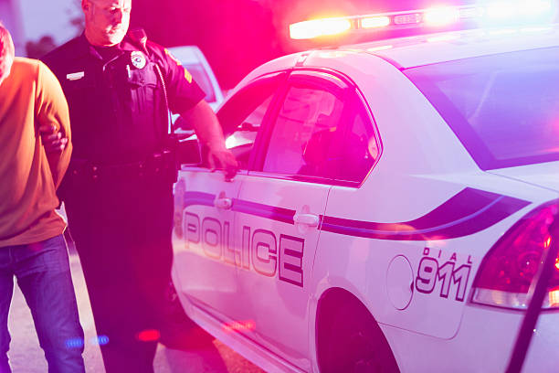 Police officer arresting a young man at night Police officer making an arrest, escorting a young man in handcuffs toward the back seat of his police car, at night. police lights stock pictures, royalty-free photos & images