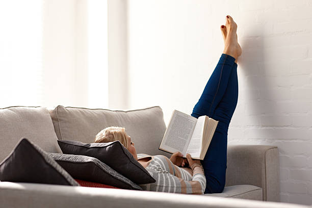 Mature woman relaxing on sofa reading a novel Mature woman relaxing on sofa reading a novel at home feet up stock pictures, royalty-free photos & images