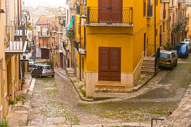 Corleone, small town in Province of Palermo in Sicily. Very narrow cobbled street with old houses and some cars in distance parked at the side. 
