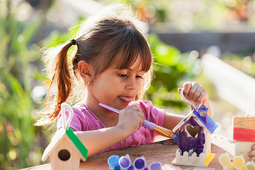 A cute little Hispanic girl with pigtails is working on an art project outdoors on a sunny day.  She is holding a paintbrush, decorating a wooden birdhouse for Earth Day.