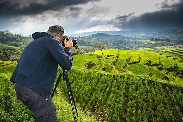 A matured man landscape photographer is looking through the viewfinder of his camera and takes photographs of beautiful terrace rice fields in Flores island, Indonesia, with a tele photo lens mounted on his tripod.