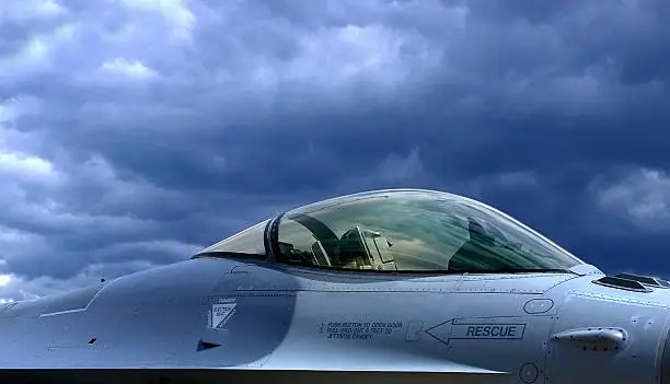 A F-16 fighter jet photographed from the side with gray clouds in the background