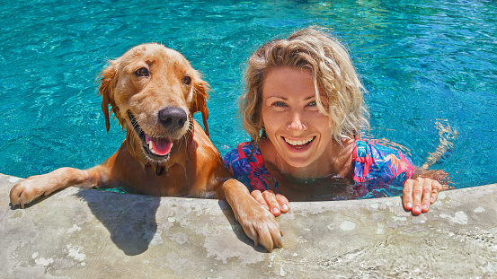 Funny portrait of smiling woman playing with dog and training golden retriever puppy in blue swimming pool. Popular dog breeds, outdoor activity and fun games with family pet on summer beach holiday.