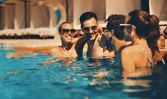 Two couples having beers while relaxing by the poolside of the hotel they stayed in. Their vacation has just started. Mid 20's men and women. Each person is wearing sunglasses.