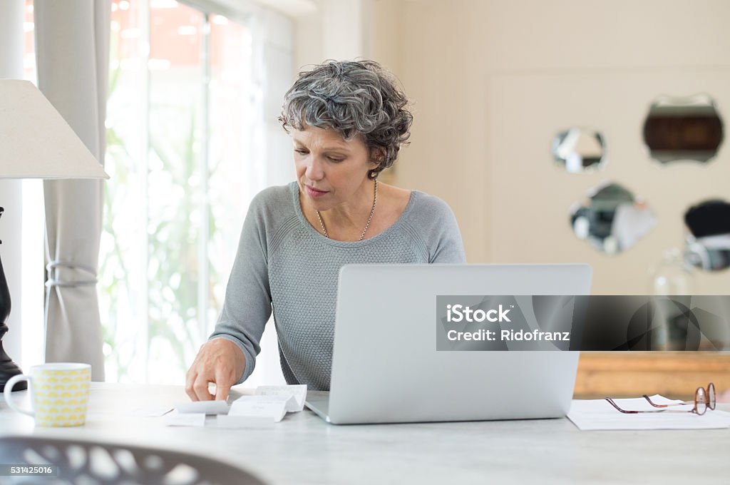 Senior woman checking bills Senior woman working on home finance. Mature woman checking bills at home. Older woman checking pension approval certificate with laptop at home. Financial Bill Stock Photo