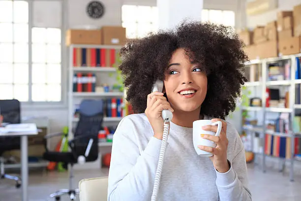 Smiling young woman drinking coffee at the office and talking on landline phone, holding a coffee cup. As background shelves with boxes and folders, desk and seats, and tall windows. Focus on foreground.