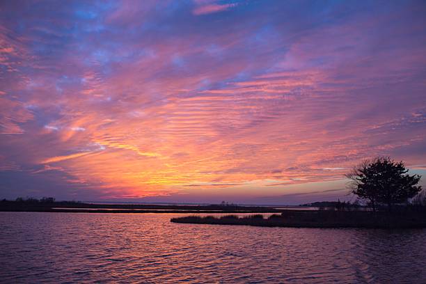 Brilliantly colorful sunset over water with rich color and reflections on the water. Taken at Assateage National Seashore at dusk, there are brilliant oranges and blues in the shot. in the distance you can see silhouetted points of land shooting in from the side of the shot.- A great coastal sunset shot taken in the outdoors.  A beautiful and Tranquil photo.