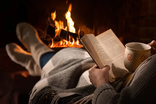 Woman resting with cup of hot drink and book near fireplace