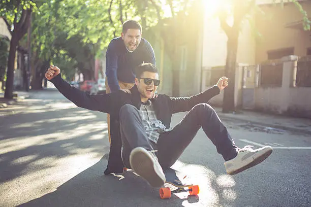 Two young men acting childish and having fun riding a longboard, pushing one another down the street.