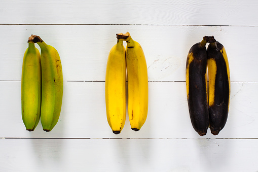 Green, yellow and black bananas arranged in a row on white wooden table.