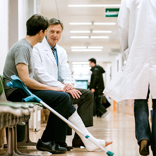 Man With Cruches And Cast On Broken Leg Consulting Doctor Mature man with broken leg, cast and crutches consulting a senior doctor. Shot in hospital corridor. walking wounded stock pictures, royalty-free photos & images