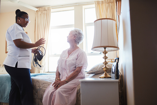 Indoors shot of home caregiver talking with senior woman sitting on bed. Female doctor visiting her senior patient at home.