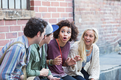 Four young multiracial adults, two men and two women, are hanging out together outside an old brick building.  An African American woman is talking, telling a story to her friends.