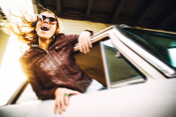 A beautiful smiling young woman hangs out the passenger side window of a moving classic car, her hair blowing in the wind.  She wears sunglasses, the summer sun shining behind her.  Horizontal image with copy space.  Some motion blur.