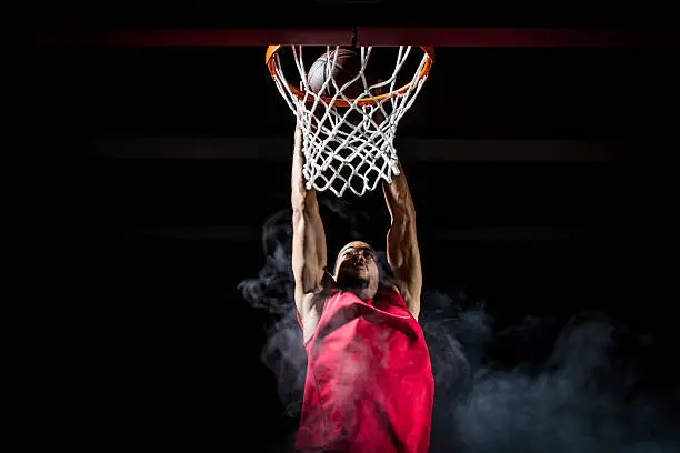 Front centre view of young basketball player in red jersey in mid-air taking a dunk shot with both of his hands. Black background with smoke.