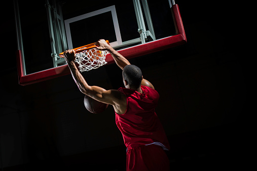 Rear-view shot of a young basketball player in a red jersey in mid-air taking a dunk shot, with both hands clothing the ring.