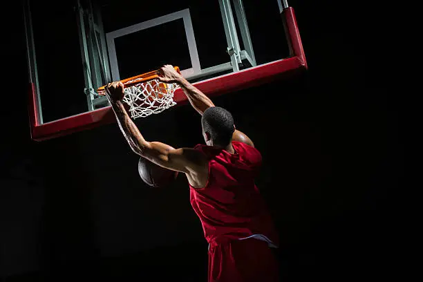 Rear-view shot of a young basketball player in a red jersey in mid-air taking a dunk shot, with both hands clothing the ring.