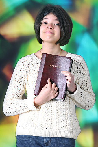 A pretty tween girl proudly displaying her Bible for the viewer in front of a large, stained glass window.