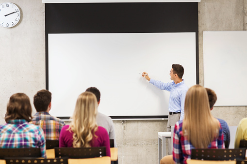 education, high school, teamwork and people concept - smiling teacher standing in front of students and writing something on white board in classroom