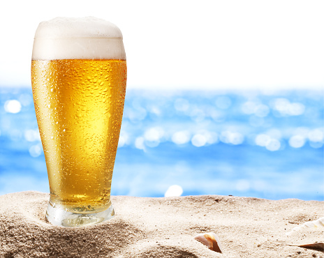 Photo of cold beer botle in the sand. Sparkling sea at the background.