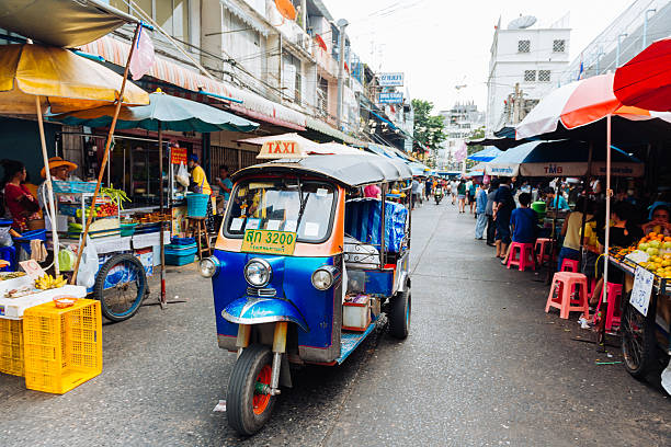 Bangkok Thailand Tuktuk Drives Silom Neighborhood Market Street Bangkok, Thailand - December 3, 2014: A small city street leading to Silom Road is lined with people shopping and selling at the fruit markets. A tuktuk taxi drives past the vendors' stalls. silom stock pictures, royalty-free photos & images