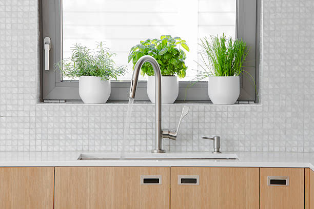 Modern kitchen Stainless steal faucet with water running into sink. Horizontal image of modern kitchen detail. Showing a kitchen sink and faucet with water running from the faucet, and potted herbs behind on window sill. Space for copy. kitchen sink stock pictures, royalty-free photos & images