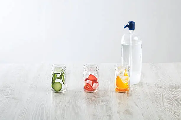 Rustic jars with ice and various fillings: orange,strawberry,cucumber and mint prepared to make fresh homemade lemonade with sparkling water from syphone Healthy beverage to cool down in summer time