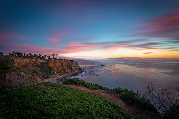 A lighthouse shines its light beam at sunset over the water with an island in the background.