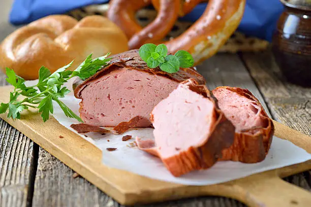 Traditional oven fresh Bavarian meat loaf with a brown crispy crust on a wooden cutting board garnished with parsley and oregano, a roll and a pretzel in the background in soft focus