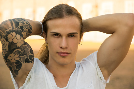 Handsome young man outdoors, with hands behind head, looking intensely at camera with confident expression