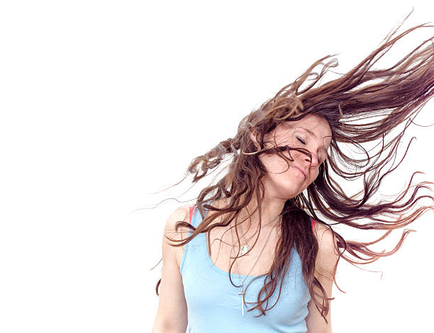 A young attractive female with strawberry blonde hair has her arms crossed as she leans into a strong wind which is blowing her hair completely to the side - isolated on a blue background. Canon 5D MarkII.