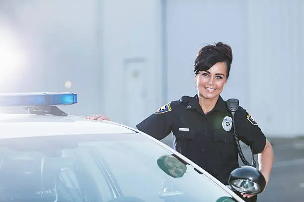 A policewoman standing next to her patrol car, smiling and looking at the camera with her hands on her hips. She is wearing the standard navy blue policeuniform.