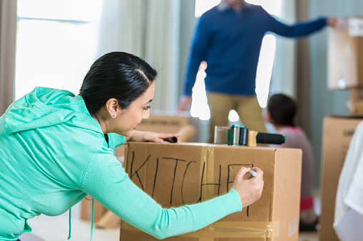 Mid adult Hispanic woman writes 'kitchen' with a marker. She has just packed up some of the kitchen supplies as she and her family prepare to move. Her husband and daughter are working in the background.