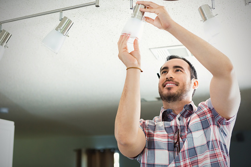 Hispanic man reaches up to install or change light bulb on light fixture in new hoe. He is standing on a ladder and reaching up to the track lighting fixture. He smiles confidently as he works. He is wearing a plaid shirt and has brown hair and a beard.