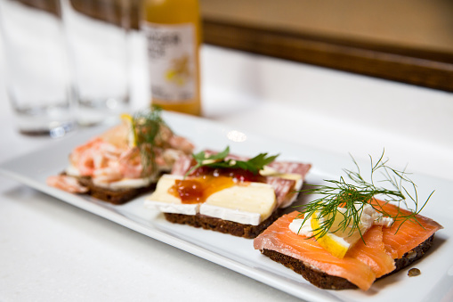 A close up of three different types of open sandwiches on a white rectangular plate. Salmon with lemon and dill in foreground focus with the background in bokeh. 
