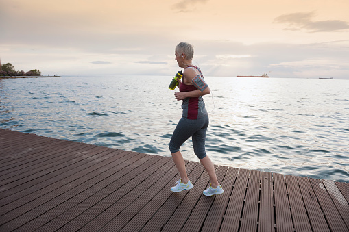 Mature-senior woman jogging at the coast, she using mobile phone and drinking water
