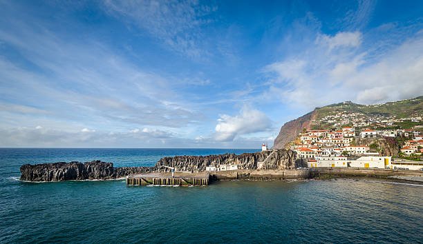 Panoramic view of Camara de Lobos fishing village harbor. Old town buildings, pier and small lighthouse on the rocky shore of Atlantic ocean. Madeira island, Portugal.