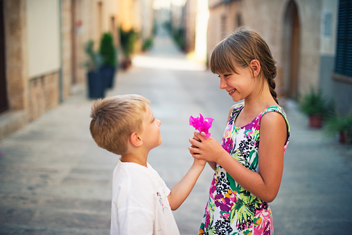 Little boy and girl looking in each other's eyes. The boy aged 4 is giving a flower to a girl aged 8. Mediterranean town street.
